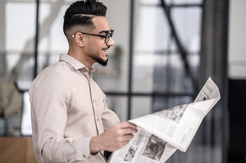 Interest. Smiling young bearded man in glasses standing sideways to camera reading newspaper with interest indoors - Foto, afbeelding