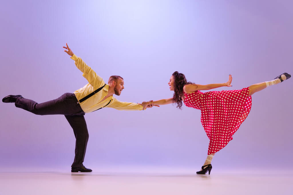 Lindy Hop. Active and emotional couple in colorful retro style costumes dancing incendiary dances isolated on purple background in neon. Actors in motion and action. Concept of art, 60s, 70s culture - Fotografie, Obrázek