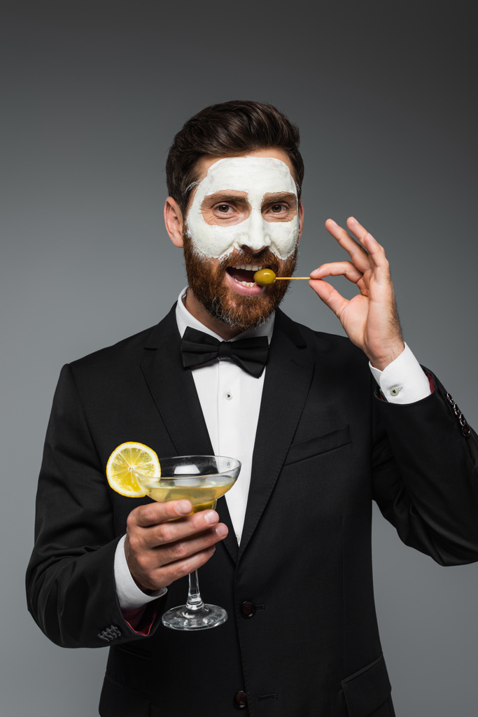 γενειοφόρος άνδρας με κοστούμι με μάσκα από πηλό στο πρόσωπο που κρατά κοκτέιλ και τρώει ελιά που απομονώνεται σε γκρι  - Φωτογραφία, εικόνα