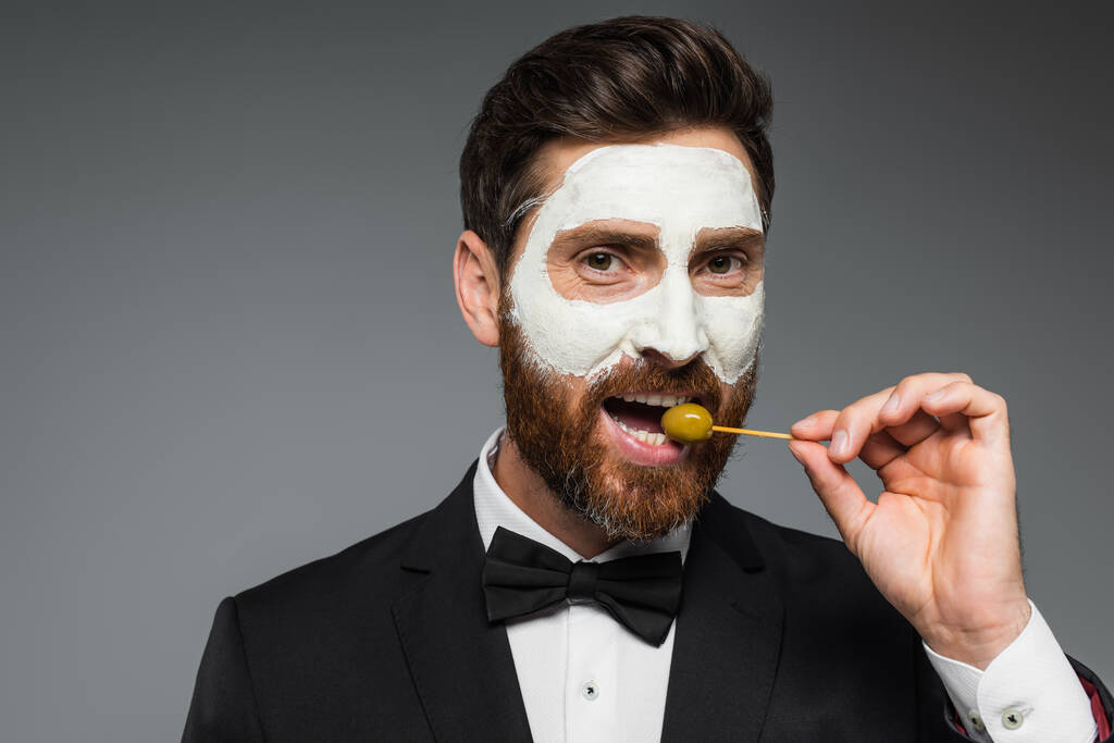 bearded man in suit with clay mask on face holding toothpick and eating olive isolated on grey - Zdjęcie, obraz