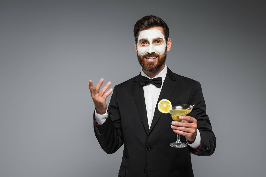 joyful bearded man in suit with clay mask on face holding cocktail isolated on grey - Foto, afbeelding