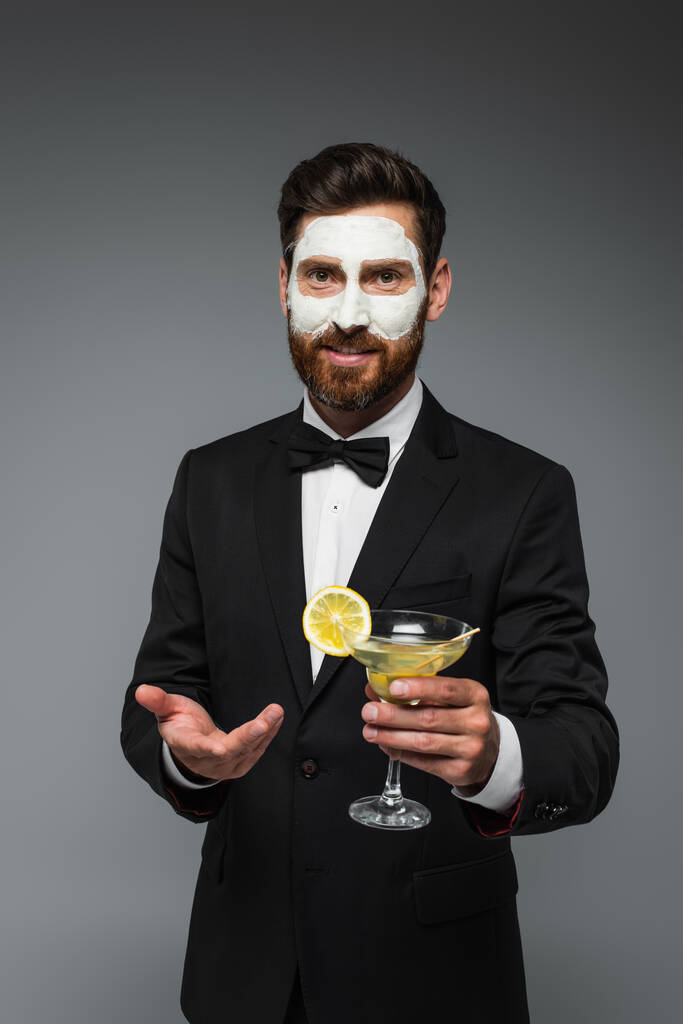 bearded man in tuxedo with clay mask on face holding cocktail and gesturing isolated on grey - Foto, imagen