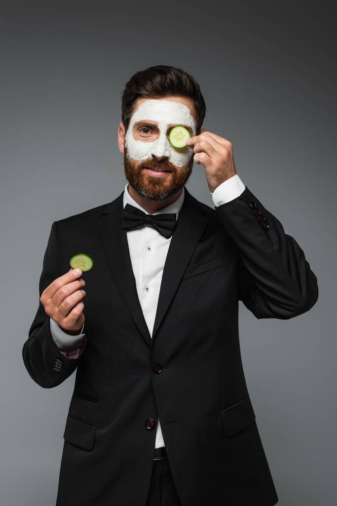 бородатый мужчина в костюме с глиняной маской на лице, держащий кусочки огурца изолированные на сером  - Фото, изображение
