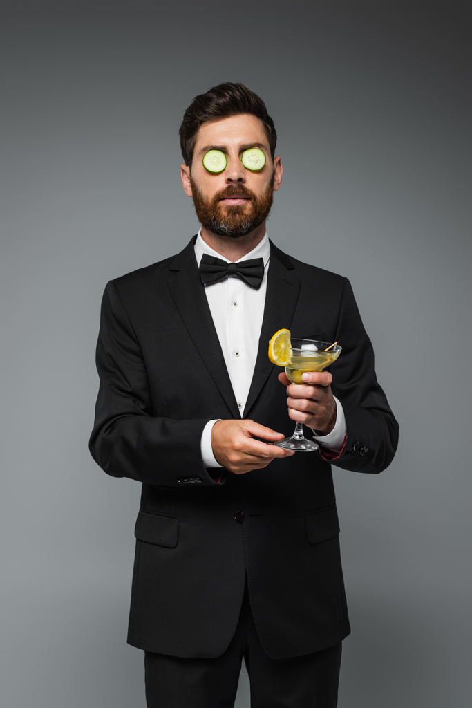 bearded man in tuxedo with fresh sliced cucumber on eyes holding glass of cocktail isolated on grey - Photo, image