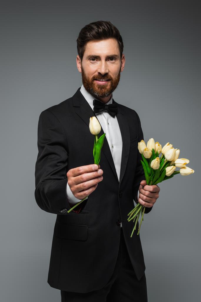 бородатий чоловік в елегантному тукседо з краваткою з бантом тримає тюльпани ізольовані на сірому
  - Фото, зображення