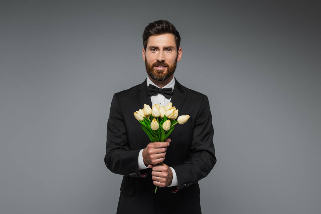 bearded man in elegant suit with bow tie holding bouquet of tulips isolated on grey  - Zdjęcie, obraz