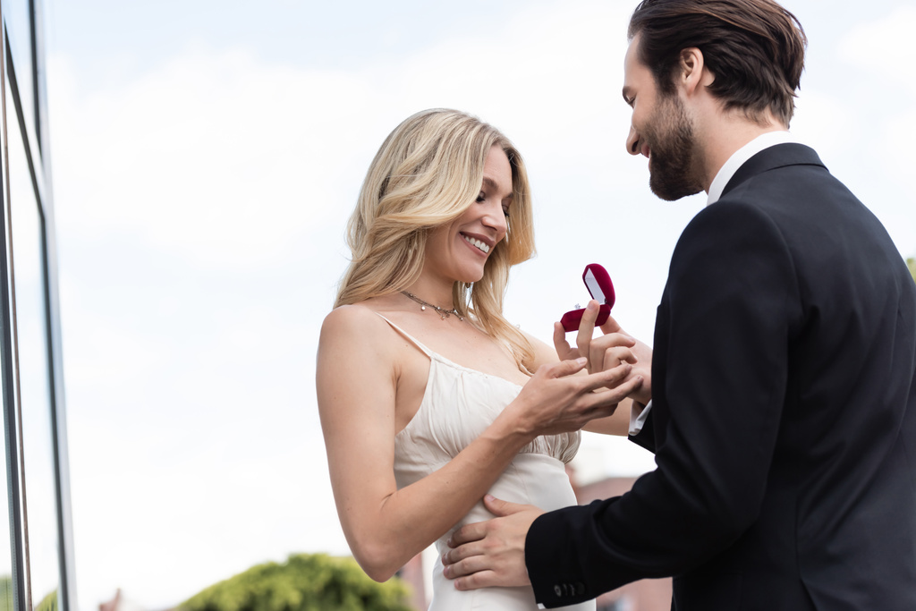 Low angle view of smiling man holding engagement ring near girlfriend in dress on terrace  - Fotoğraf, Görsel