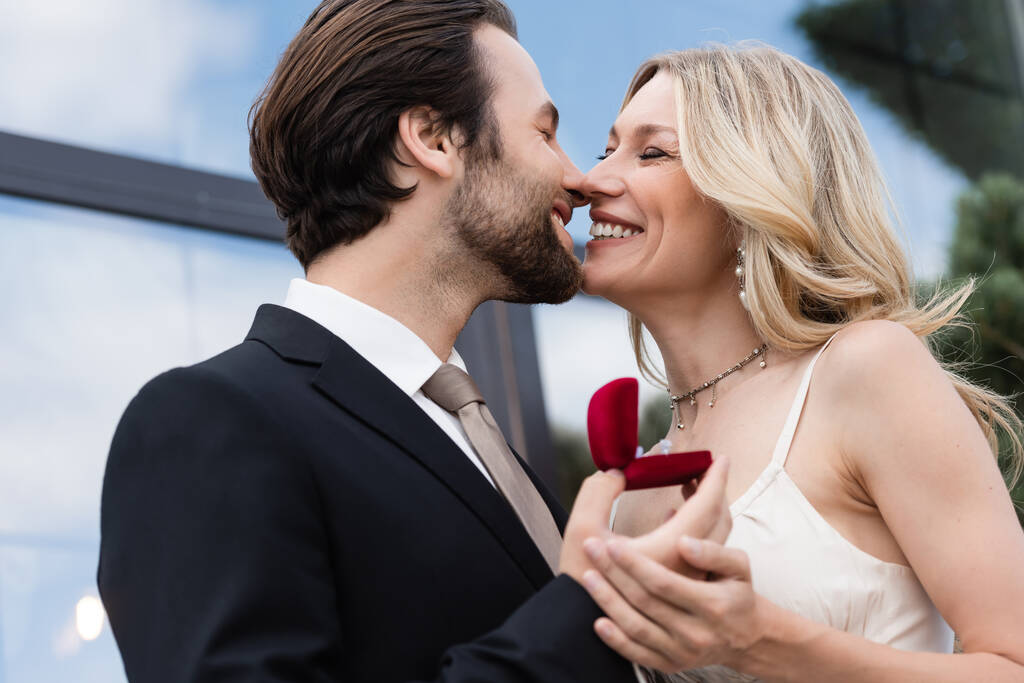 Smiling man in suit holding engagement ring and kissing girlfriend on terrace  - Fotoğraf, Görsel