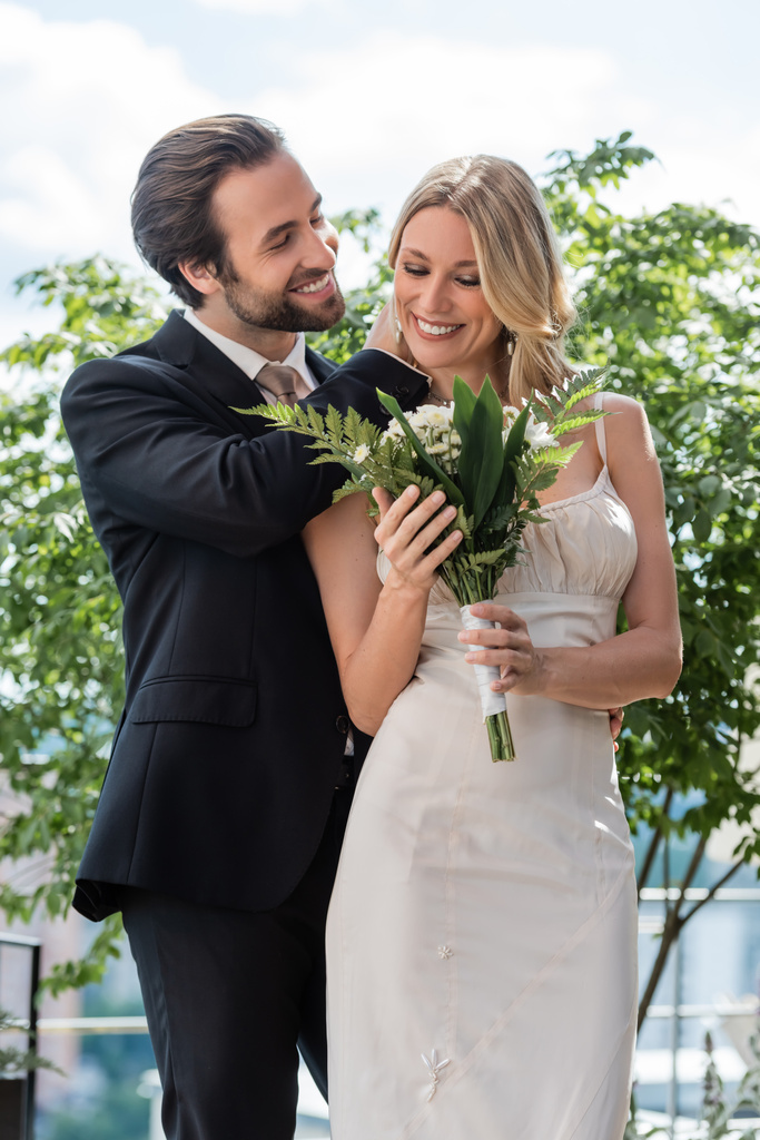 Smiling groom touching bride with bouquet on terrace of restaurant  - Photo, Image