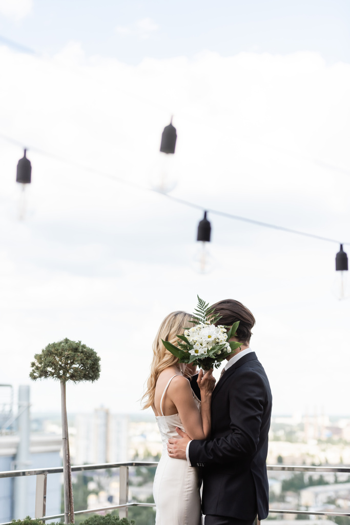 Newlyweds covering faces with bouquet while kissing on terrace  - Foto, afbeelding