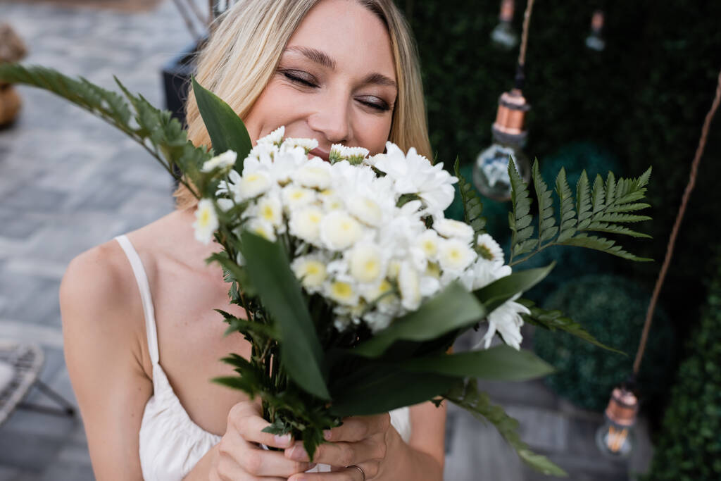 Blonde bride covering face with blurred bouquet on terrace  - Foto, Imagen