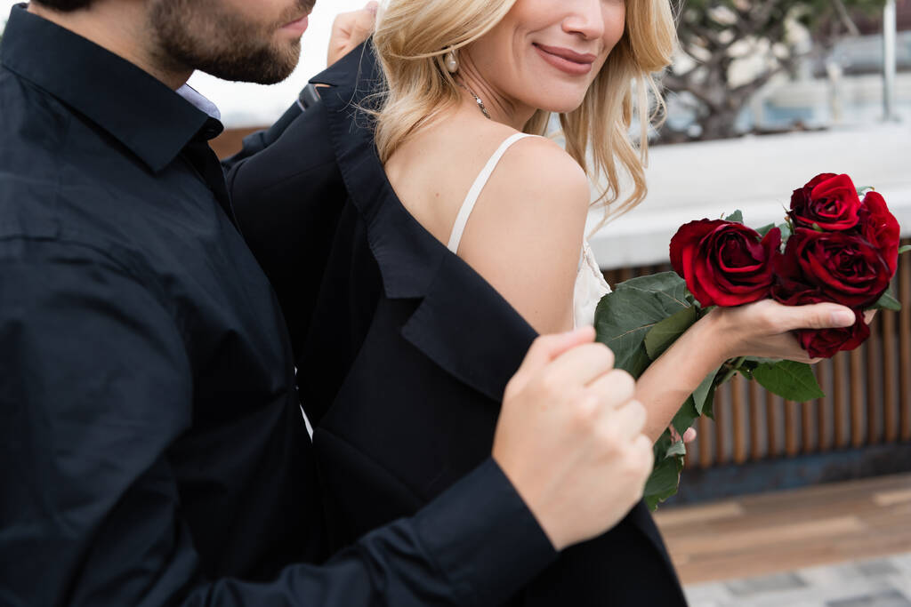Cropped view of man wearing jacket on blonde girlfriend with roses outdoors  - Zdjęcie, obraz