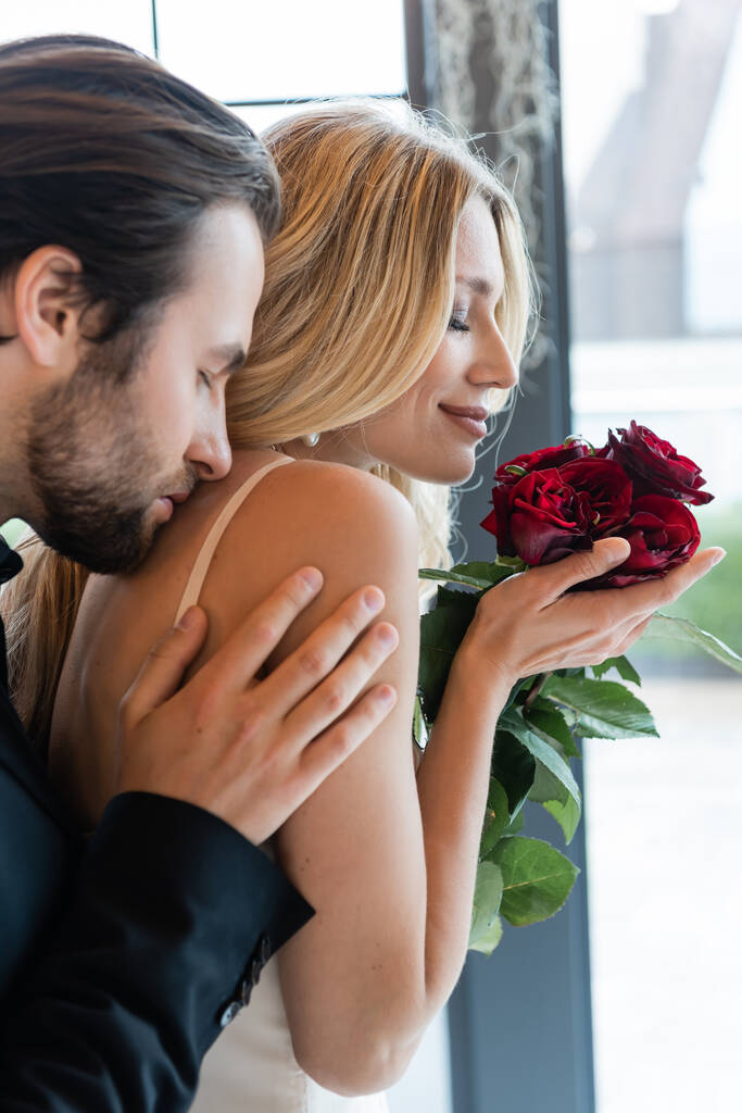 Вид збоку на брюнетку чоловік цілує усміхнену дівчину з трояндами в ресторані
  - Фото, зображення