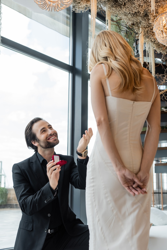 Positive man holding engagement ring near girlfriend in dress in restaurant  - Zdjęcie, obraz