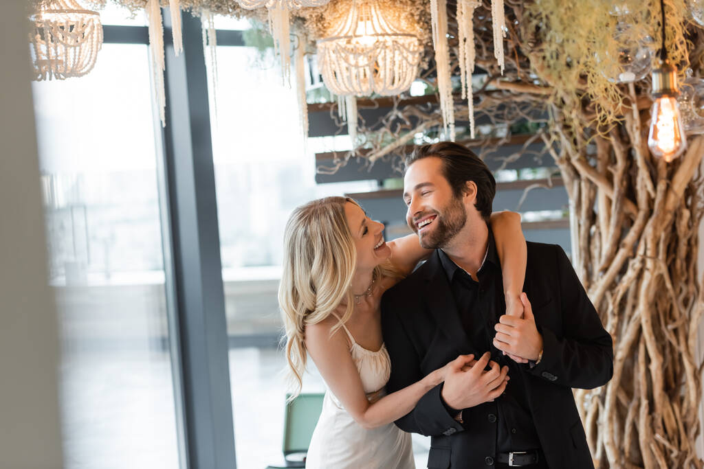 Cheerful woman in dress holding hands of boyfriend under chandelier in restaurant  - Fotoğraf, Görsel
