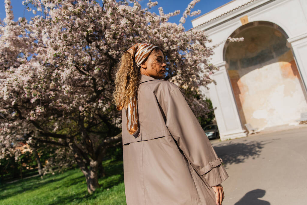 молодая африканская американка в платке и стильном плаще, стоящем рядом с цветущим деревом в Праге - Фото, изображение