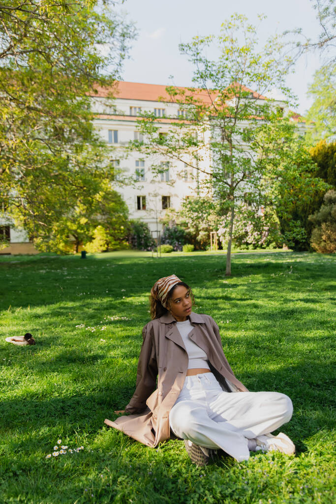 young african american woman in headscarf and stylish trench coat sitting on green lawn in park - Foto, imagen
