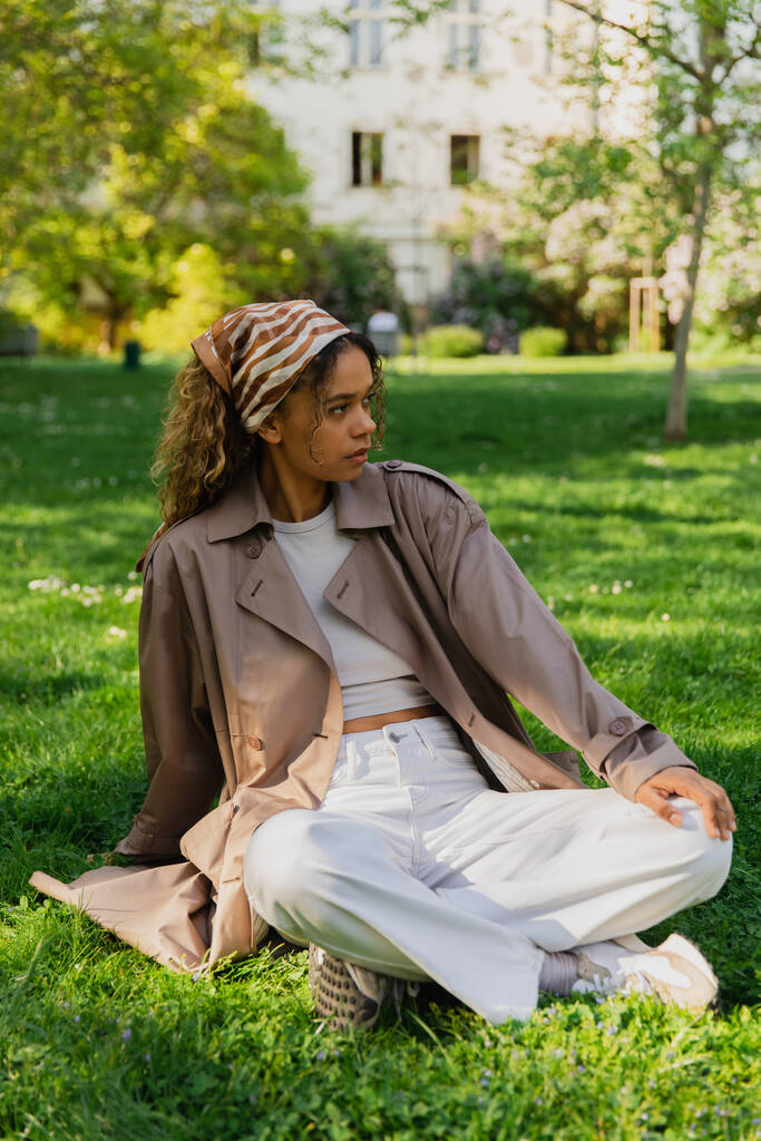 stylish african american woman in headscarf and trench coat sitting on green lawn in park - Fotoğraf, Görsel