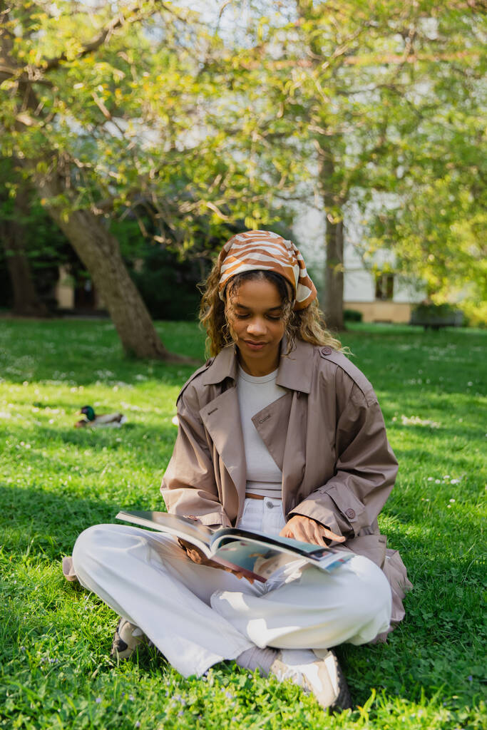 Αφροαμερικανή γυναίκα με μαντίλα και καμπαρντίνα κάθεται στο πράσινο γκαζόν και διαβάζοντας περιοδικό στο πάρκο - Φωτογραφία, εικόνα