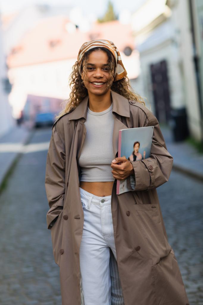 щаслива афроамериканська жінка в хедкарфі і траншеї пальто, що йде з журналом на вулиці в празі
  - Фото, зображення