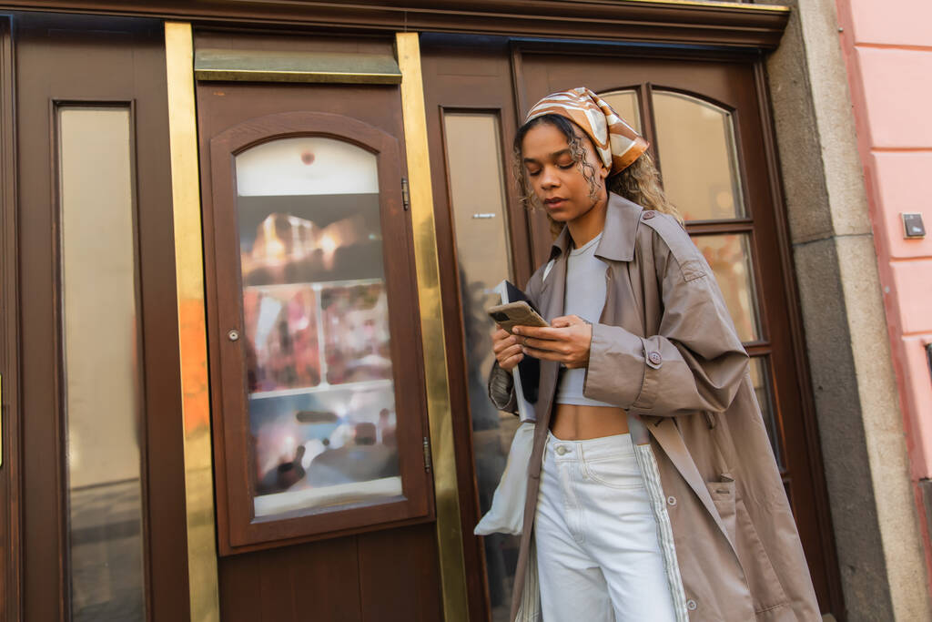 stylish african american woman in headscarf and trench coat walking and using smartphone in prague  - Foto, immagini