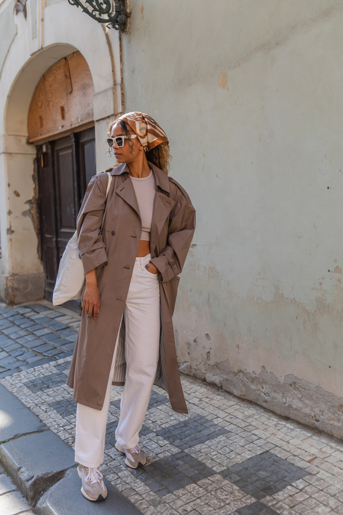 Полная длина стильная африканская американка в платке и солнцезащитных очках позирует с многоразовой сумкой на улице в Праге  - Фото, изображение