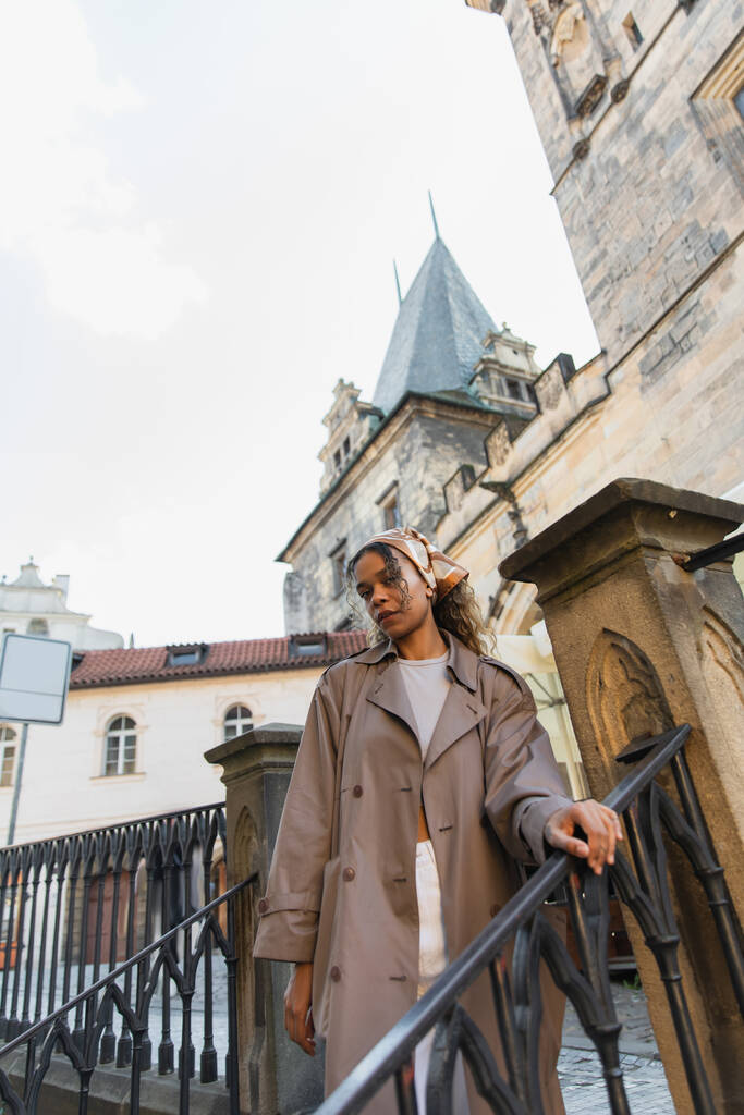 low angle view of stylish african american woman in trench coat near old town hall tower in prague - Foto, imagen