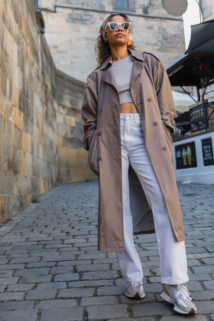 full length of stylish african american woman in trendy trench coat and sunglasses posing on street in prague - Foto, immagini