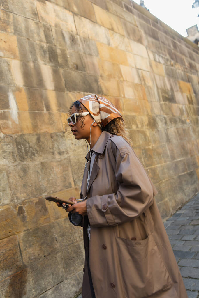 πλαϊνή άποψη της αφρικανικής αμερικανικής γυναίκας σε μοντέρνα αξεσουάρ ακούγοντας μουσική σε ενσύρματα ακουστικά και κρατώντας smartphone  - Φωτογραφία, εικόνα