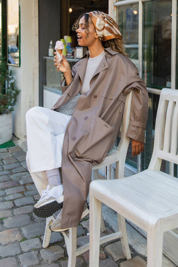 amazed african american woman in headscarf and stylish trench coat holding ice cream cone and sitting on chair outside - 写真・画像