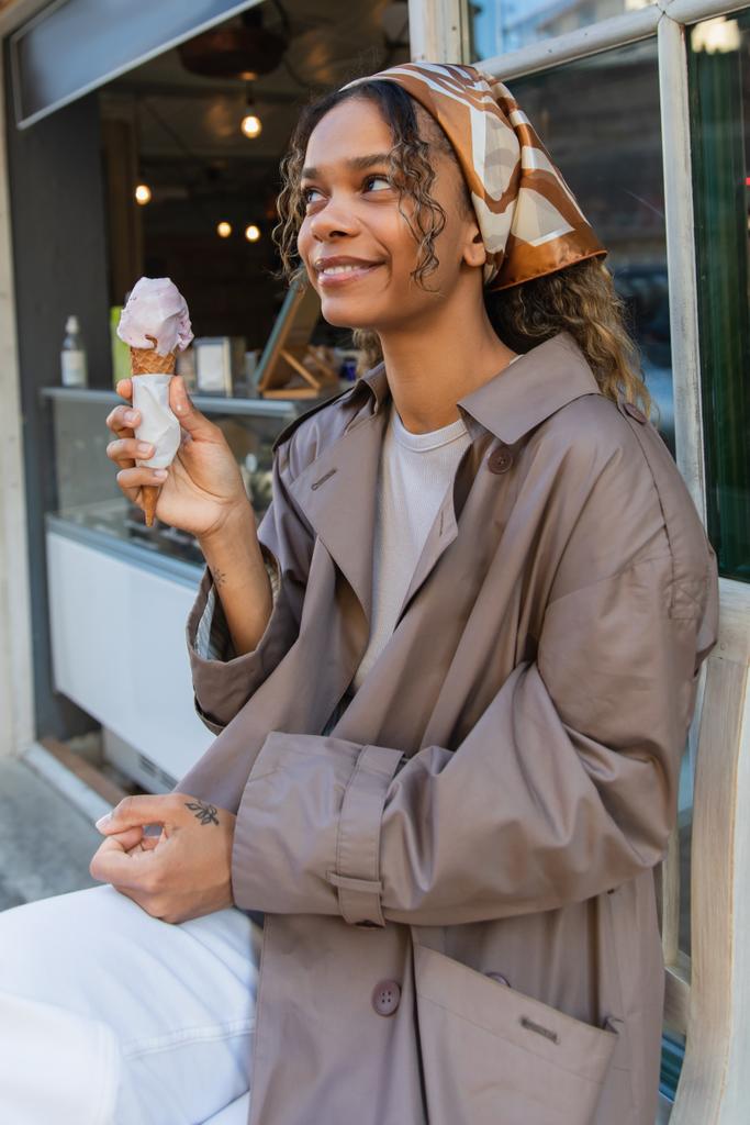 cheerful african american woman in headscarf holding ice cream cone and sitting on chair in prague - Foto, imagen