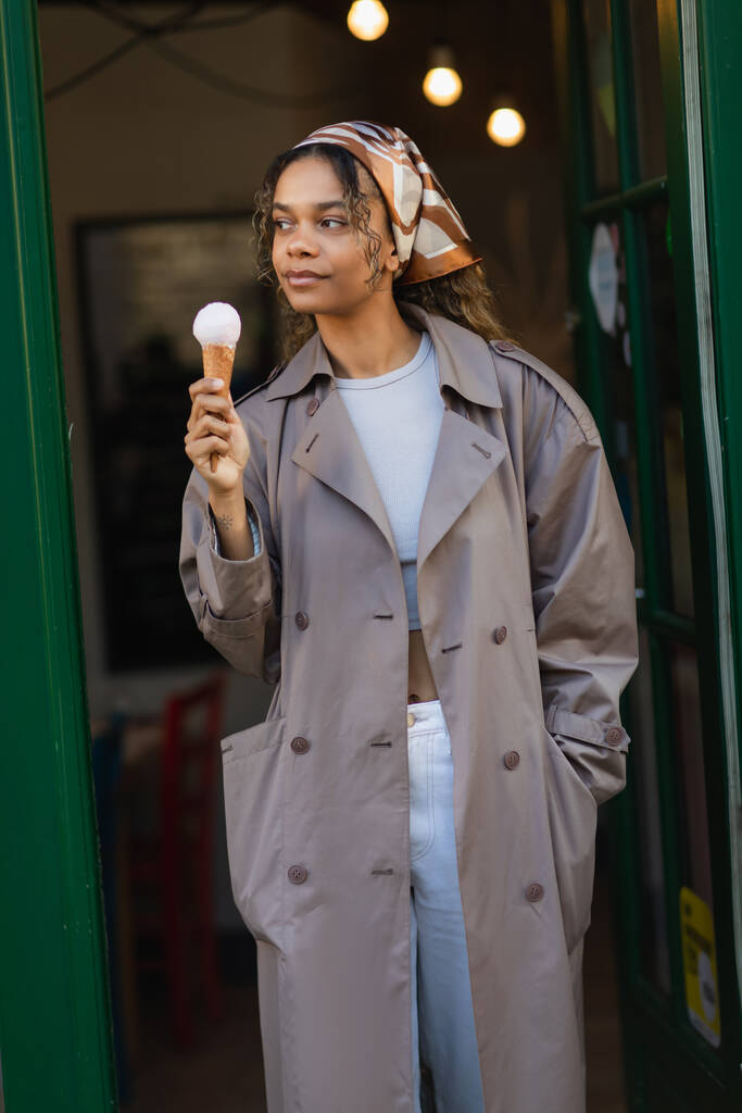 молодая африканская американка в платке и стильном плаще держит мороженое на улице в Праге  - Фото, изображение