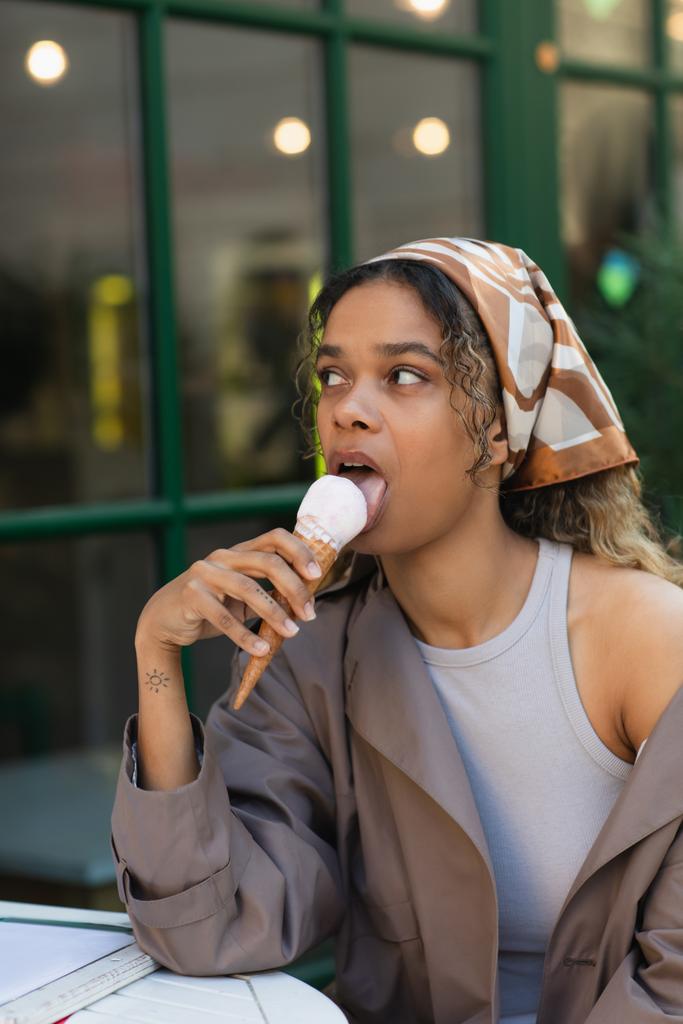 tattooed african american woman in headscarf and stylish trench coat eating ice cream and sitting in cafe terrace  - Foto, Bild