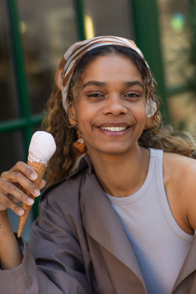 happy african american woman in headscarf holding ice cream cone and sitting in cafe terrace  - Foto, Imagem