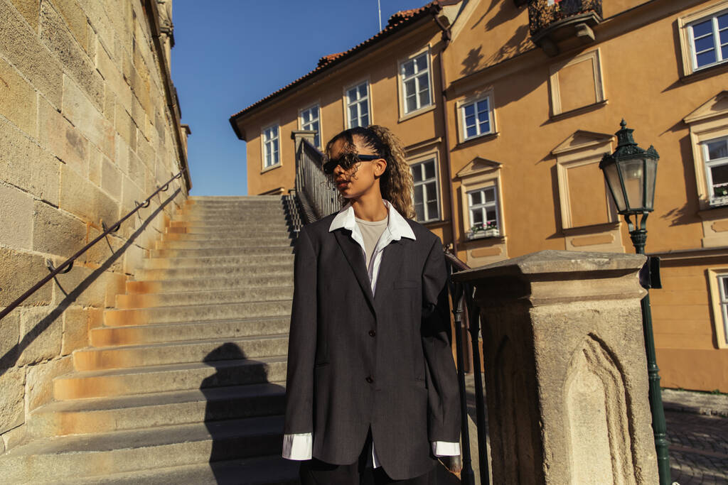 stylish african american woman in sunglasses and oversize suit standing near stairs on street in prague  - Foto, afbeelding