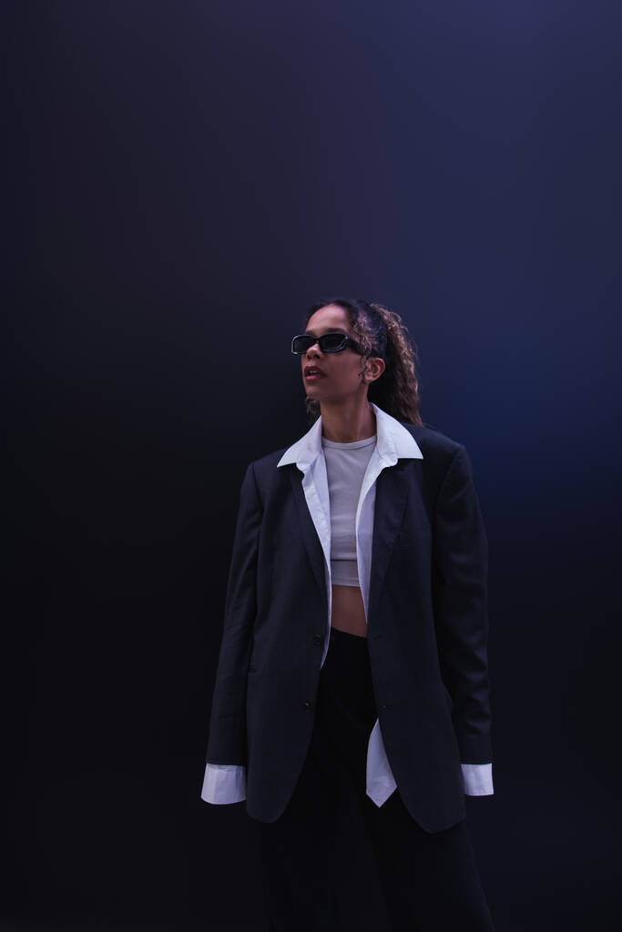 stylish african american woman in sunglasses and oversize suit on dark blue - 写真・画像