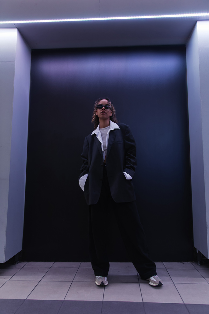 stylish african american woman in sunglasses and oversize suit posing with hands in pockets near dark blue wall - Foto, immagini