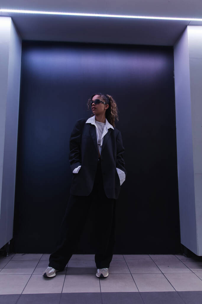 full length of african american woman in sunglasses and oversize suit posing with hands in pockets near dark blue wall - Fotoğraf, Görsel
