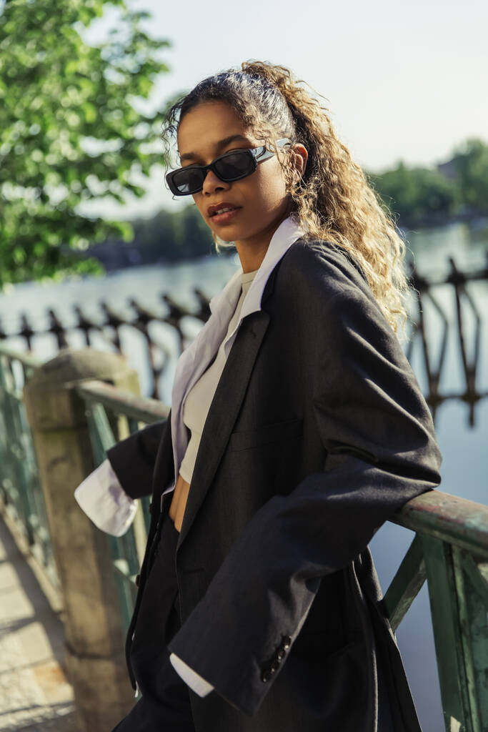 curly african american woman in oversize suit and stylish sunglasses standing near river in prague  - Fotografie, Obrázek