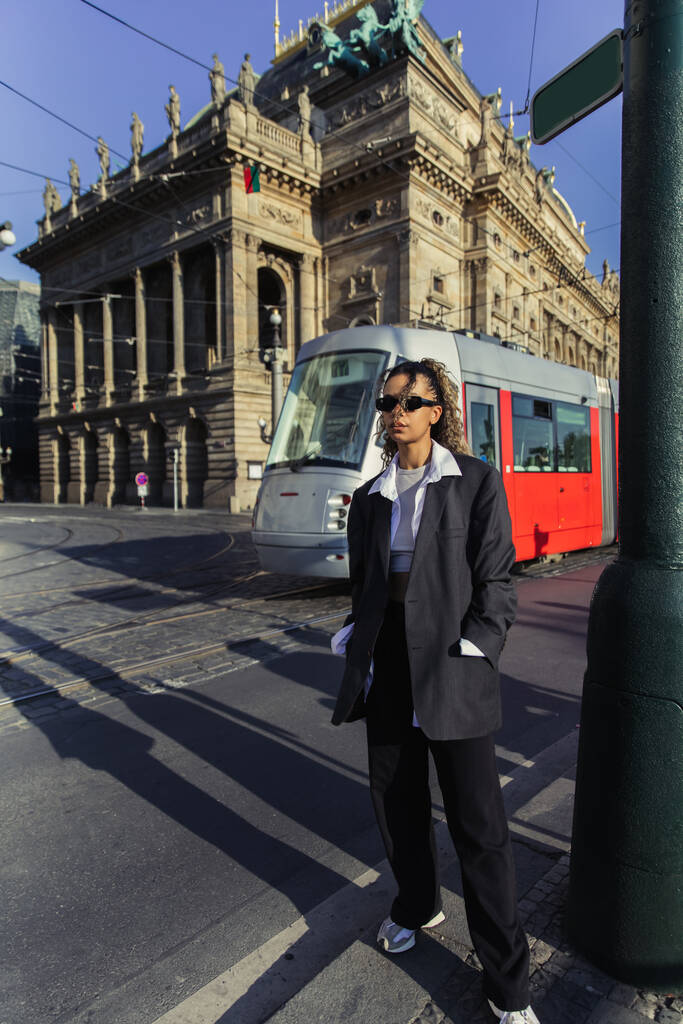stylish african american woman in oversized blazer and sunglasses standing with hands in pockets near national theatre in prague - Photo, image