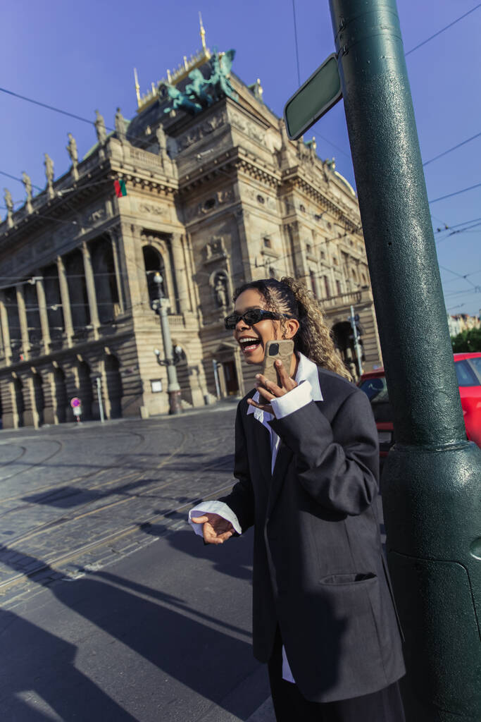 happy african american woman in oversized blazer and sunglasses holding smartphone near national theatre in prague - 写真・画像