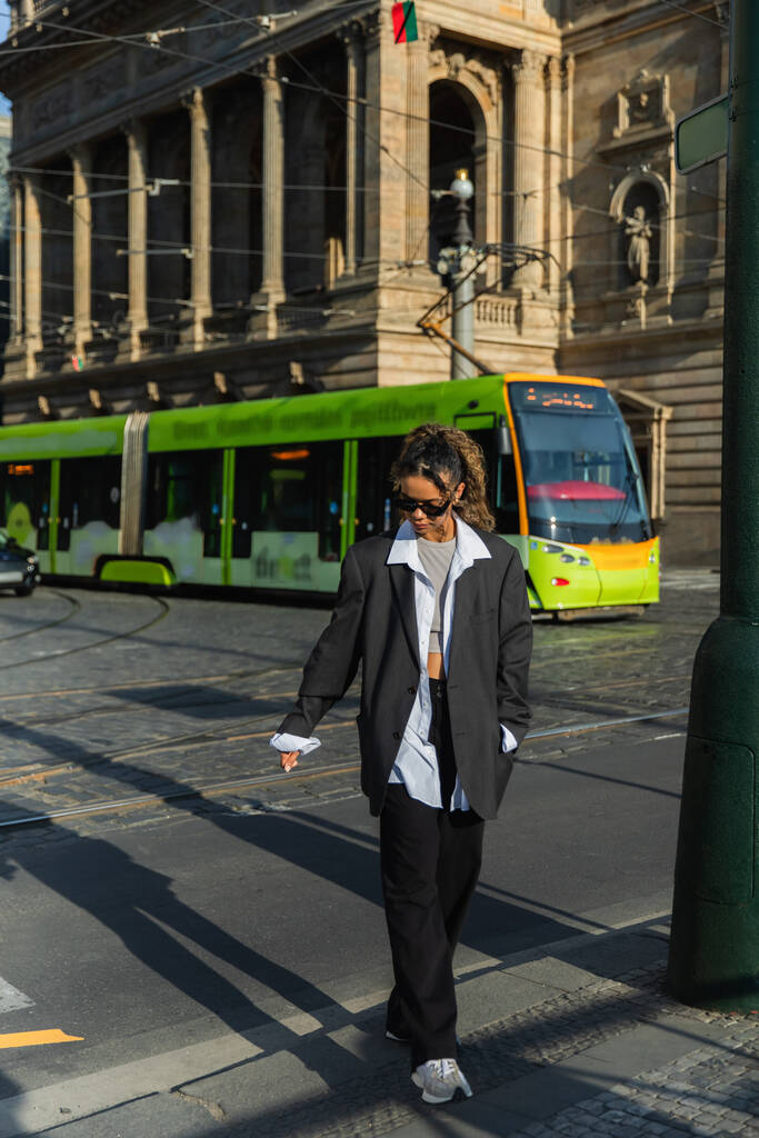 young african american woman in stylish blazer and sunglasses walking with hand in pocket near tram and building in prague - Photo, Image