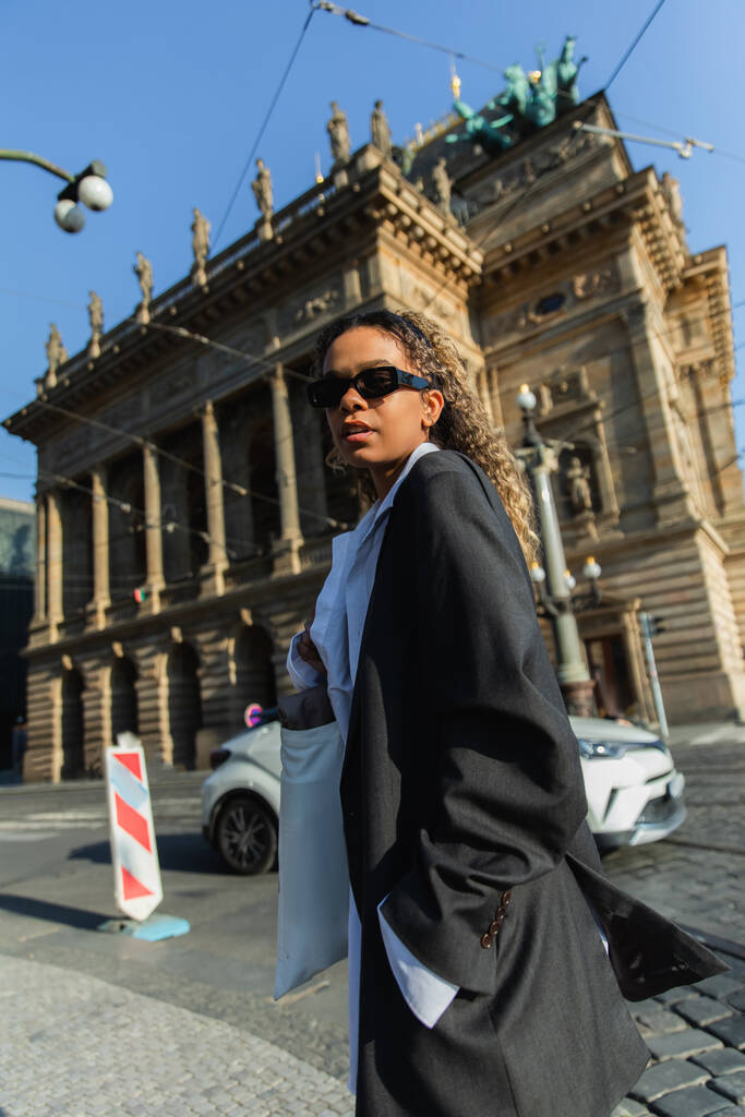 low angle view of young african american woman in stylish blazer and sunglasses near national theatre in prague - Fotografie, Obrázek