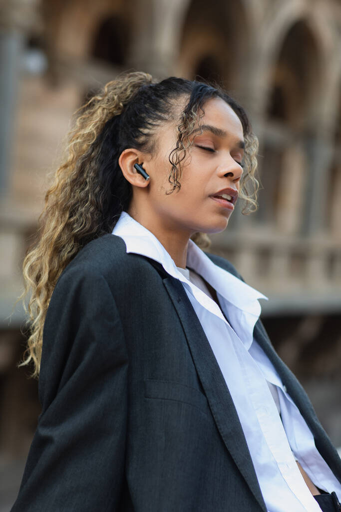 african american woman with closed eyes listening music in wireless earphone on urban street of prague  - Foto, Bild