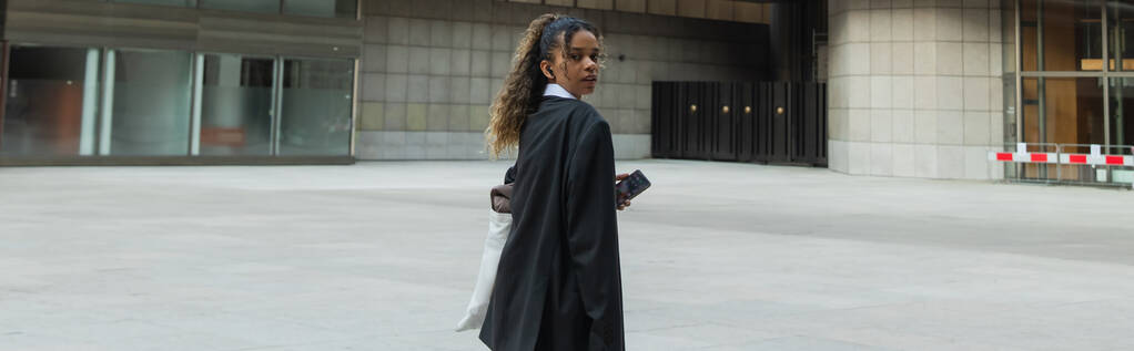 african american woman in oversize suit listening music in wireless earphone and walking on urban street in prague, banner - Fotografie, Obrázek