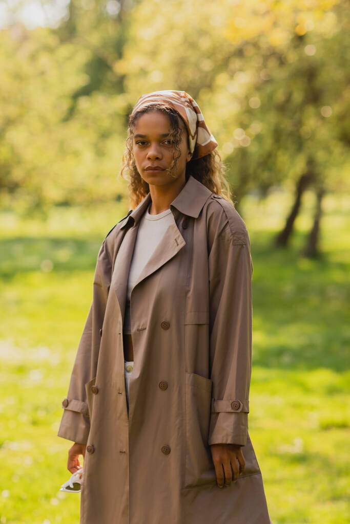 young african american in headscarf and beige trench coat standing in park - Fotó, kép