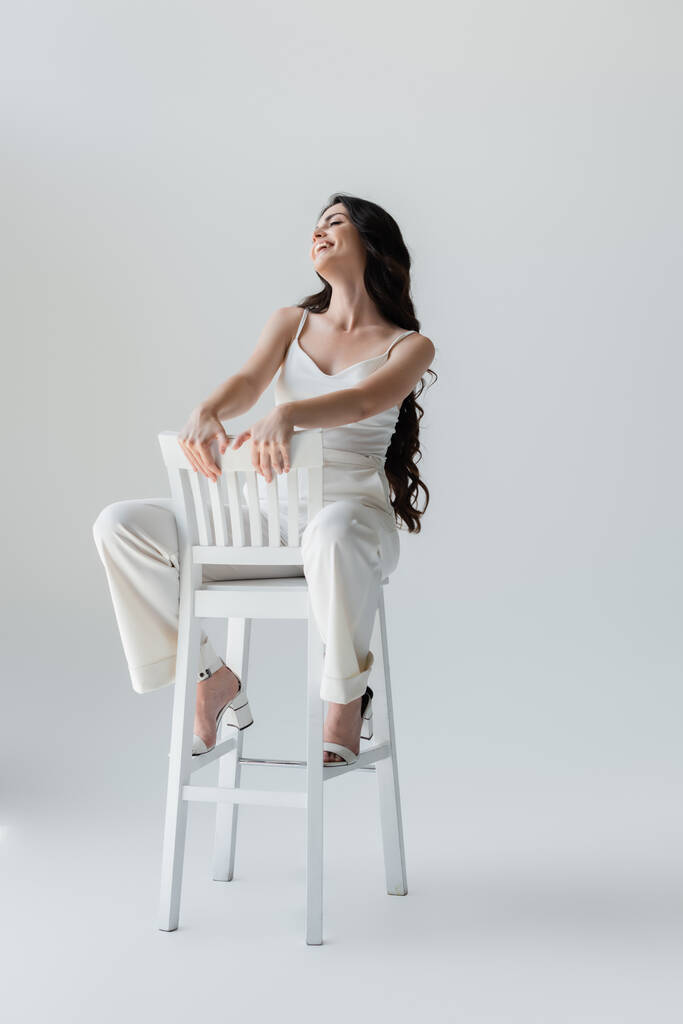 Full length of stylish brunette woman smiling while sitting on chair on grey background - Foto, Imagem