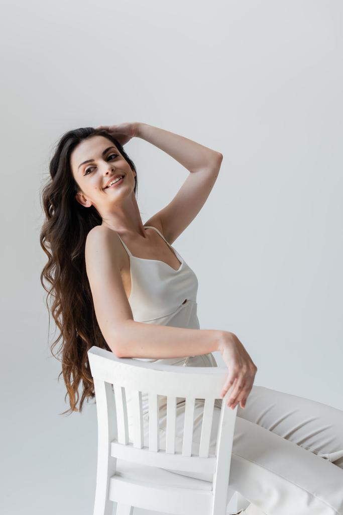 Positive long haired woman sitting on white chair isolated on grey  - Foto, Imagen