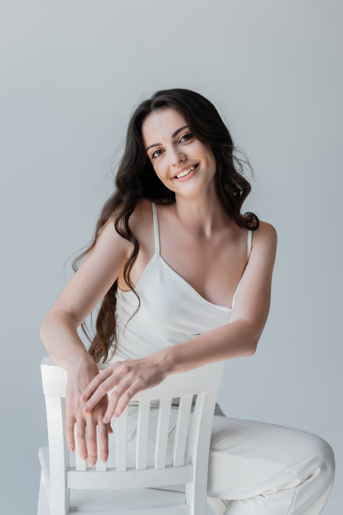 Happy brunette woman looking at camera while posing on chair isolated on grey  - Fotoğraf, Görsel