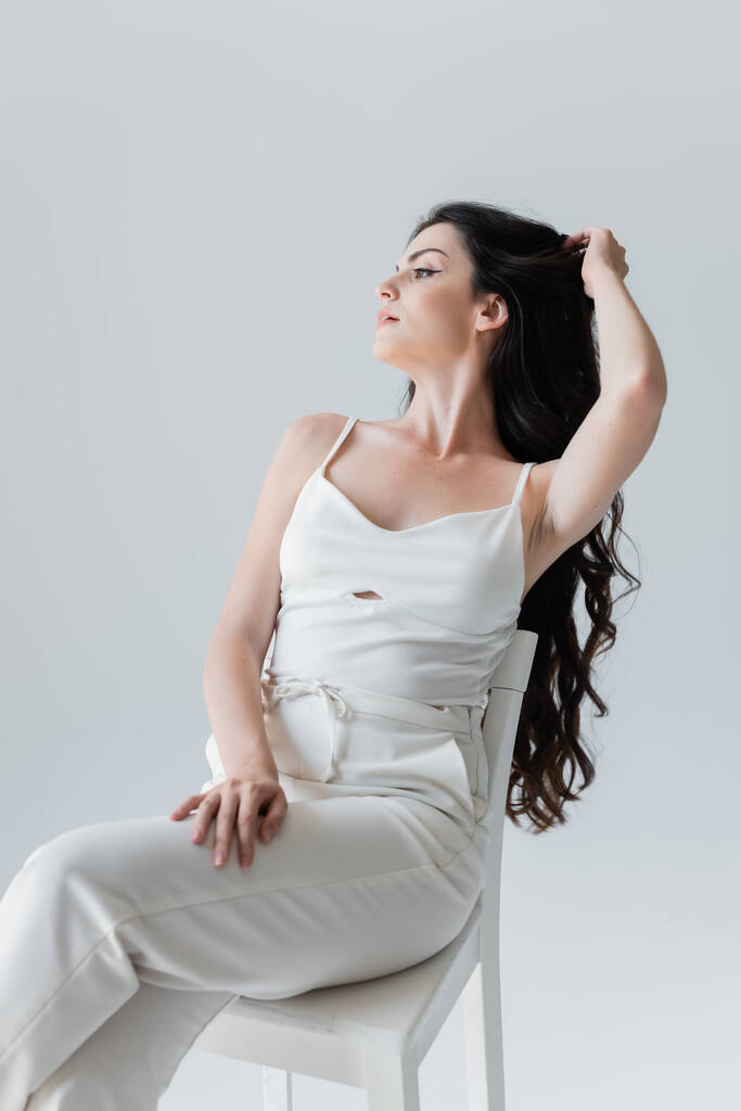 Young model in white clothes touching long hair while sitting on chair isolated on grey  - Fotografie, Obrázek
